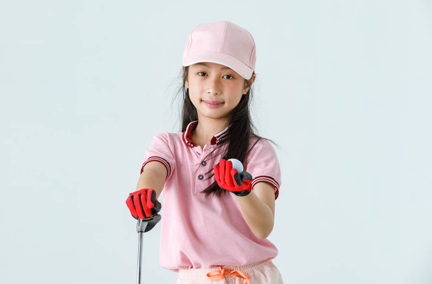 Закрыть изолированный студийный снимок мяча для гольфа в руке азиатской маленькой профессиональной девочки-гольфистки в розовой спортивной одежде красные перчатки и кепка держат палец нацеливаясь на цель на зеленой траве. - Фото, изображение