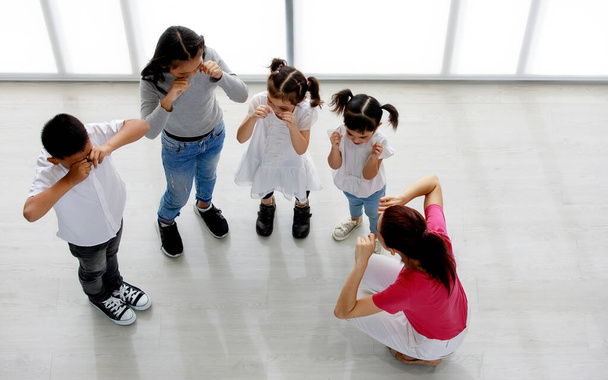 Top view felvétel ázsiai nő általános iskolai tánc tanár ül tanítani csoport kisfiú és a lányok, hogy végre sírás testtartás boldog szórakoztató magán óvoda színjátszó osztály stúdió szobában. - Fotó, kép