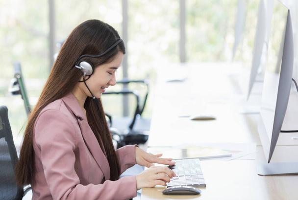 Asian young beautiful happy confident professional female operator wears pink formal suit and microphone headset sitting talking solving problem with customer while typing information with keyboard. - Foto, Imagen