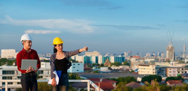 アジアの男性と女性のエンジニアが忙しいハードハットチームで、屋根の上に立って、青い空を背景にした屋外での撮影を指摘しながらプロジェクトに取り組んでいます. - 写真・画像