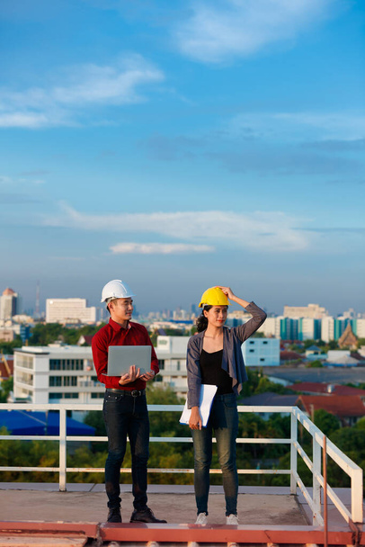 Занятые азиатские инженеры мужского и женского пола в команде хардхатов, работающие вместе, стоя на крыше и работая над проектом, указывая в сторону, открытый снимок с голубым небом. - Фото, изображение