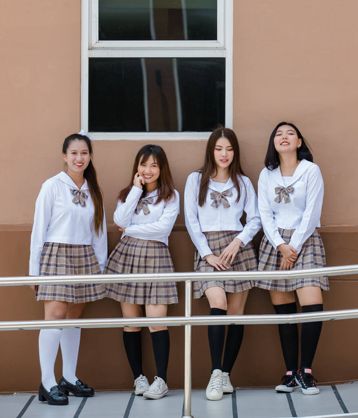 Група з чотирьох молодих привабливих азіатських студентів середньої школи в коричневій картатій шкільній формі, що стоїть разом у шкільному кампусі посміхаючись. Концепція освіти та життя старшокласників
. - Фото, зображення