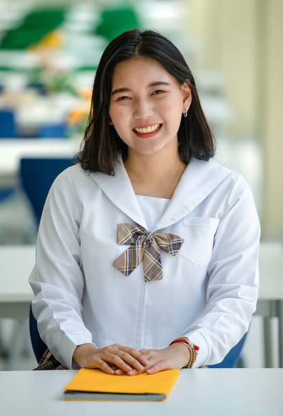 白いシャツ茶色の若い魅力的な女性のアジアの高校生の肖像画は、学校のキャンパス内の教室に座って学校の制服をチェックしました。教育・高校生生活のコンセプト. - 写真・画像
