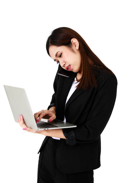 Ασιάτης νεαρός μακριά καστανά μαλλιά αρκετά επιτυχημένη γραμματέας επιχειρηματίας φοράει μαύρο επίσημο κοστούμι κρατήσει γκρι φορητό υπολογιστή στο χέρι σταθεί χαμόγελο ματιά στην οθόνη κατά την πληκτρολόγηση μπροστά από το λευκό φόντο. - Φωτογραφία, εικόνα