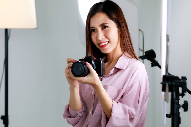 Asian beautiful adult female photographer with long hair wearing casual shirt, holding a camera and taking photo in indoor studio with blur background of  lighting equipments - Foto, Bild