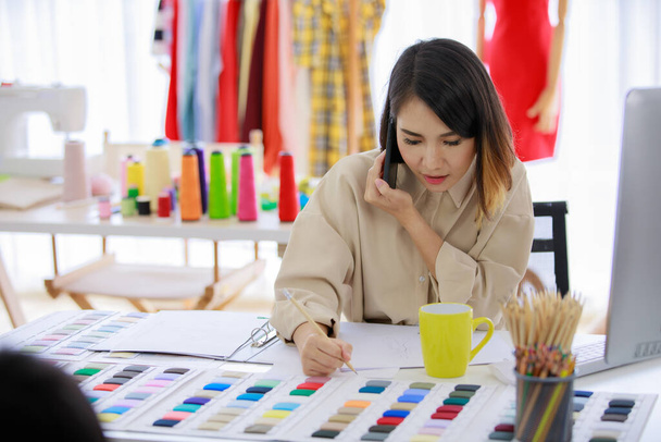 Азійська жінка тримає смартфон, розмовляючи з клієнтом. Мода дизайнерського одягу в магазині тканин Tailor. Концепція клієнта професії дизайнера Fashion. - Фото, зображення