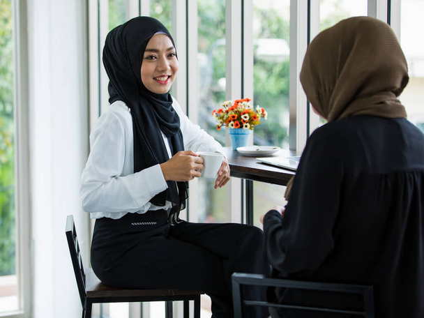 Veselé mladé ženy ze Středního východu v tradičních šátcích, popíjejí kávu a přátelsky si povídají u stolu s laptopem během přestávky na moderním pracovišti - Fotografie, Obrázek