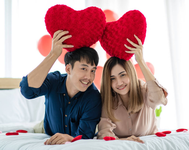 Pozytywny azjatycki mężczyzna i kobieta patrząc w aparat z uśmiechem i demonstrując poduszki w kształcie serca podczas odpoczynku na łóżku podczas obchodów Walentynek. Miłość i relacje nastolatków kochanków koncepcja. - Zdjęcie, obraz