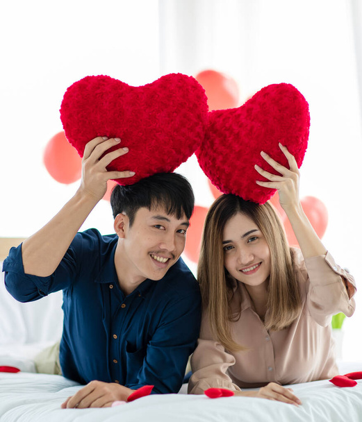 Homme et femme asiatiques positifs regardant la caméra avec le sourire et montrant des oreillers en forme de cœur tout en se reposant sur le lit pendant la célébration de la Saint-Valentin. Amour et relation des amants adolescents concept. - Photo, image