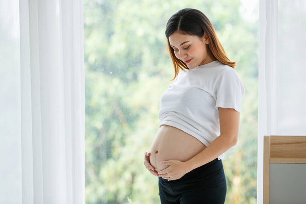 Portrait of a good-looking Asian pregnant woman dress casually standing and touching her belly with love and happiness while looking and smiling at a camera. - Photo, Image