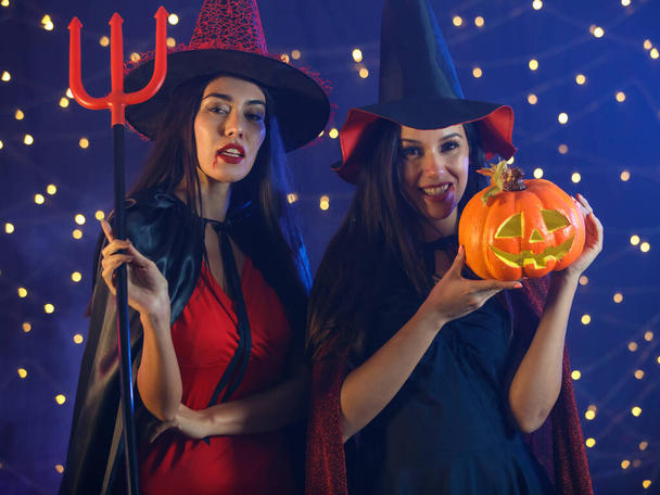 Jovens namoradas em roupas de bruxa e chapéus segurando diabo tridente e cabeça de abóbora e olhando para a câmera durante a festa de Halloween - Foto, Imagem