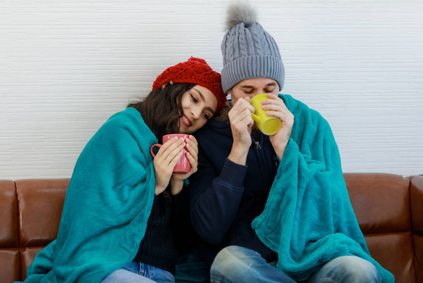 Счастливые кавказские любовники пара в шерстяной куртке свитер вязаная шляпа сидеть улыбка разговор на диване покрытия одеялом вместе держать горячий напиток кружку в руках в холодную погоду зимой. - Фото, изображение