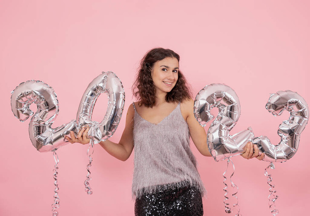 Belle fille brune aux cheveux bouclés et aux vêtements de fête posant sur un fond rose avec des confettis et tenant des ballons argentés des numéros 2023. - Photo, image