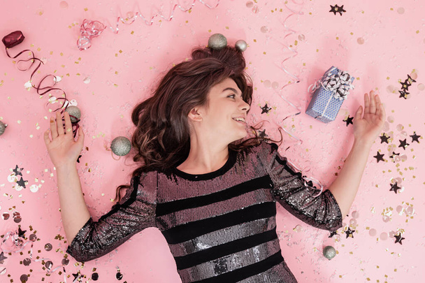 Cheerful brunette girl lies on a pink background among confetti and streamer, the concept of celebrating christmas and new year, festive party. - Фото, зображення