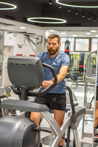 Ein aktiver Mann mit Bart trainiert im Fitnessstudio und fährt auf einem stationären Heimtrainer. Sport und Quarantäne. - Foto, Bild