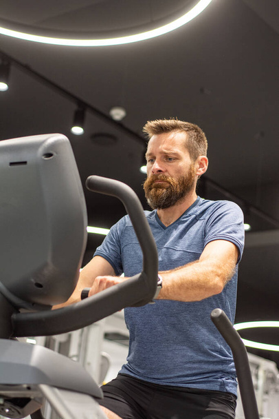 An active man with beard training in gym, riding a stationary exercise bike. Sport and quarantine. - Foto, Bild
