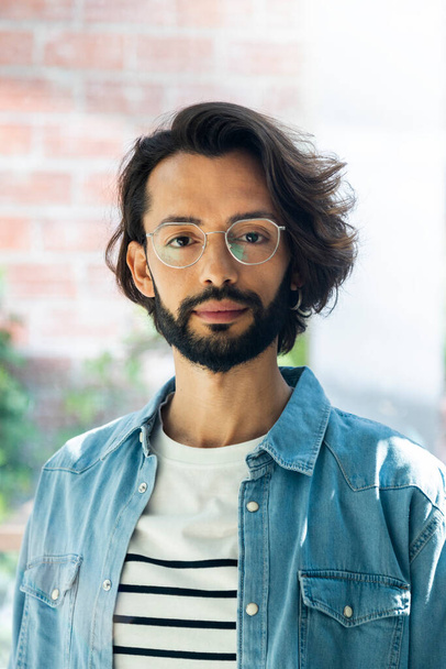 Headshot of young adult man looking at camera with dark hair and beard wearing glasses and jean shirt. Vertical image. - Фото, зображення