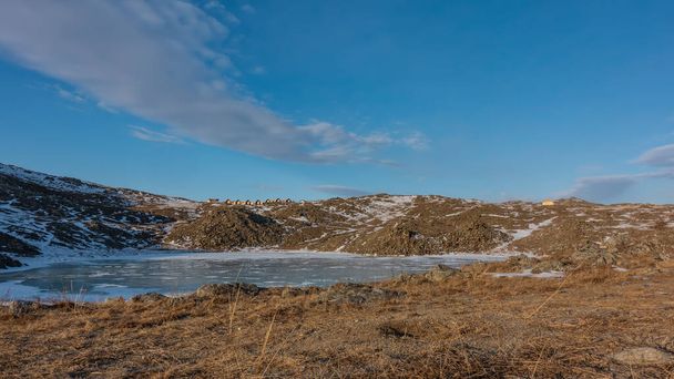 Ein kleiner zugefrorener See ist von Hügeln umgeben. Schnee auf Eis und Boden. In der Ferne, vor blauem Himmel - die Häuser der Touristenbasis. Schöne Wolken. Sibirien. Insel Olchon - Foto, Bild