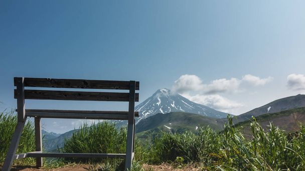 Na szczycie wzgórza, wśród zielonej trawy, znajduje się drewniana ławka. Przed nami, pod błękitnym niebem, znajduje się stożkowy wulkan z pokrytymi śniegiem zboczami. Chmury na górze. Kamczatka. Stratowulkan Vilyuchinsky - Zdjęcie, obraz