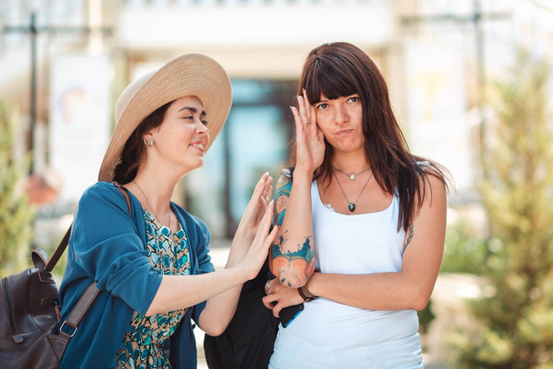 Deux jeunes femmes bavardent dans la rue. Une femme ferme sa main loin de l'interlocuteur ennuyeux. Le concept d'amitié, de communication et de psychologie. - Photo, image