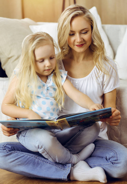 Happy family. Blonde young mother reading a book to her cute daughter while sitting at wooden floor in sunny room. Motherhood concept - Zdjęcie, obraz