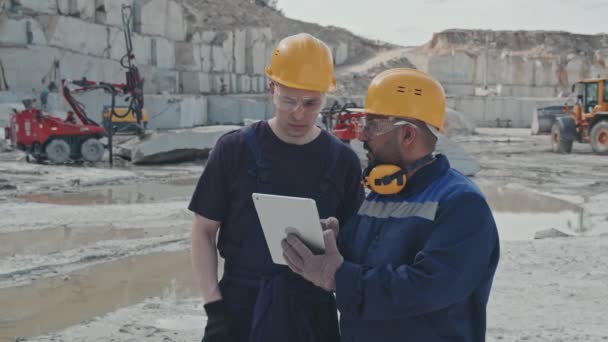 Slowmo záběr mužských dělníků v uniformách, bezpečnostních brýlích a tvrdých kloboucích při pohledu na tablet a diskusi o práci v žulovém lomu - Záběry, video