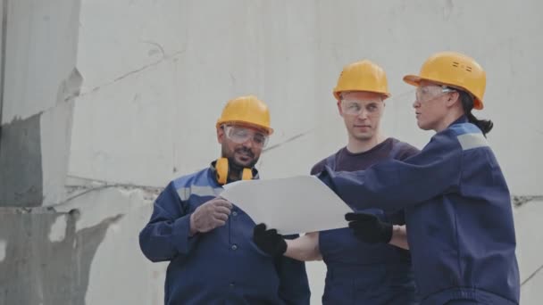 Slowmo mittlere Aufnahme von männlichen und weiblichen Arbeitern in Uniform und harten Hüten, die Dokumente oder Entwürfe im Steinbruch diskutieren - Filmmaterial, Video