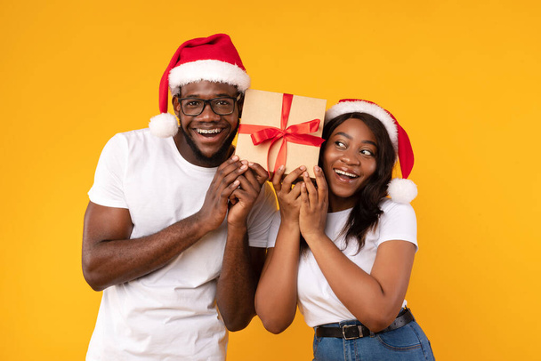 Μαύρο ζευγάρι κρατώντας χριστουγεννιάτικο δώρο μαντέψουν τι είναι μέσα, κίτρινο φόντο - Φωτογραφία, εικόνα