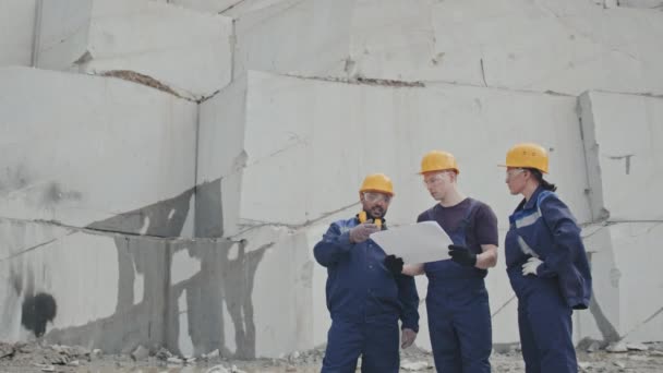 Medium shot of male and female workers in uniform, hard hats and safety goggles looking at documents or blueprints and having conversation in granite quarry - Footage, Video