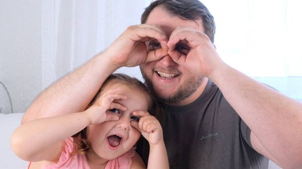 Mała córeczka 3 lata i ojciec bawią się w domu, robiąc śmieszne miny, robiąc okulary palcami, jak okulary patrzące w kamerę przez lornetkę. baw się z dzieckiem, za darmo - Zdjęcie, obraz