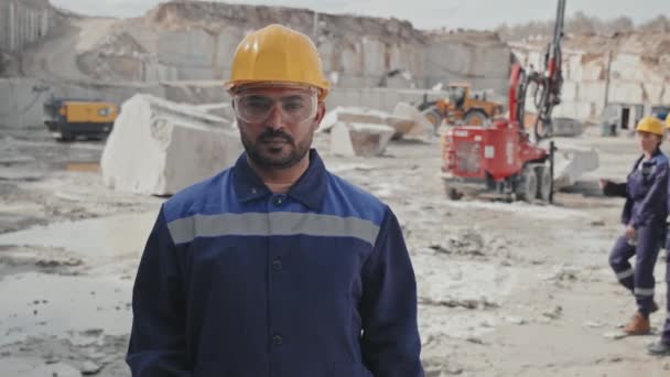 Slowmo śledzenia portret męskiego pracownika w mundurze, okulary ochronne i twardy kapelusz krzyżując ramiona i pozowanie do kamery w granitowym kamieniołomie - Materiał filmowy, wideo