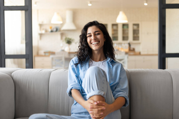 Πορτρέτο της όμορφης νεαρής μελαχρινής γυναίκας χαμογελώντας και κοιτάζοντας την κάμερα, κάθεται σε couhc στο σπίτι - Φωτογραφία, εικόνα