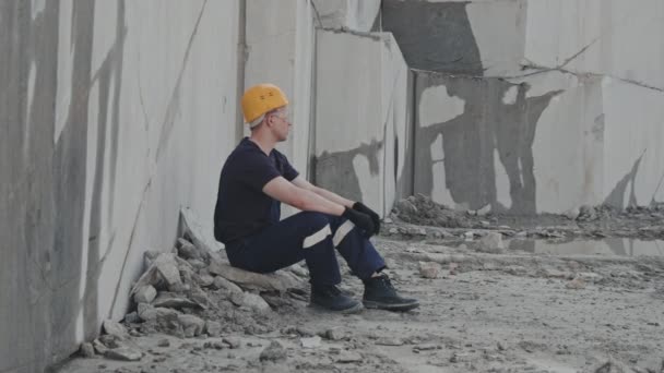 Slowmo colpo medio di lavoratore maschile stanco in cappello duro, occhiali di sicurezza e tute da lavoro seduto a terra in cava di granito e riposo - Filmati, video