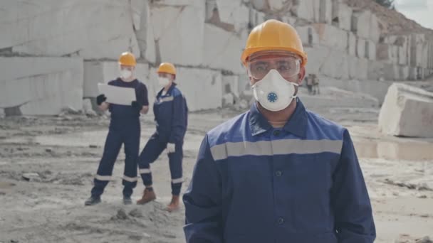 Slowmo tracking shot of male worker in uniform, gloves, dust mask, safety goggles and hard hat posing for camera in granite quarry. Male and female workers discussing documents in background - Footage, Video