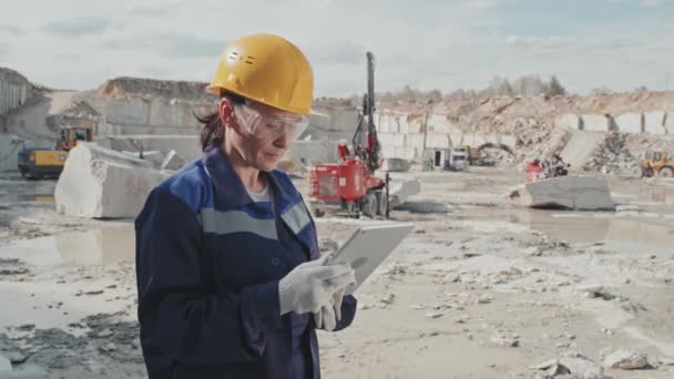 Slowmo tracking of female supervisor in uniform, safety goggles and hard hat standing in granite quarry and looking at tablet, then posing for camera. Heavy machinery and granite blocks in background - Footage, Video