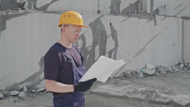 Śledzenie portretu męskiego pracownika w kombinezonach, twardych kapeluszach i okularach ochronnych stojących w kamieniołomie granitowym i czytających plany, a następnie patrzących w kamerę - Materiał filmowy, wideo