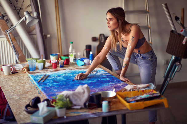 A very hot and sexy young female artist is standing over the table and thinking about a painting she is working in a relaxed atmosphere on in her studio. Art, painting, studio - Zdjęcie, obraz