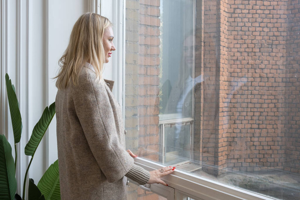Elegant blonde woman stands in front of her reflection in the window and smiles. Woman in an autumn coat looks out the window, assessing the weather. Woman dreams of the future - Photo, Image