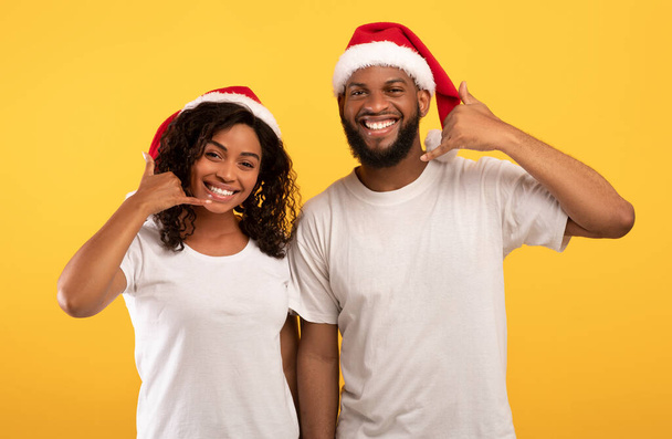 Nadšený mladý černošky pár v Santa klobouky ukazující CALL ME gesto, pózování a úsměv na fotoaparátu na žlutém pozadí - Fotografie, Obrázek
