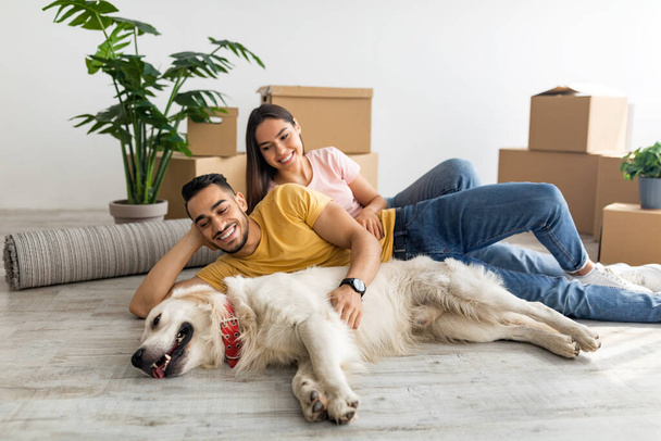 Усміхнена багаторасова пара зі своєю собакою лежить на підлозі серед картонних коробок, в приміщенні
 - Фото, зображення