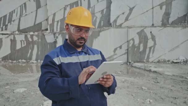 Tracking portret van Arabische man supervisor in uniform, veiligheidsbril en harde hoed met behulp van tablet en kijken naar camera in granieten steengroeve - Video