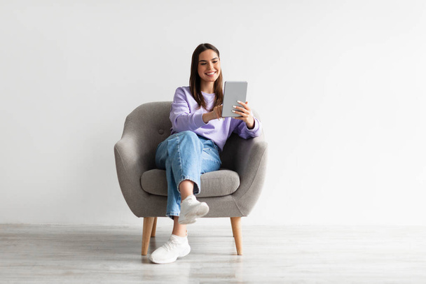 Plná délka veselá mladá žena s tabletou PC sedí v křesle, pracuje nebo studuje on-line proti bílé zdi - Fotografie, Obrázek