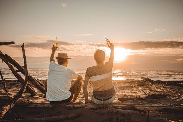 Two young friends spending time together sitting on the beach, drinking beer and toasting on vacation in twilight summer sunset. Friendship concept - Photo, Image
