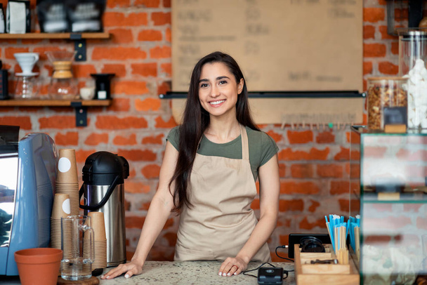 Abriendo pequeños negocios. Feliz señora latina en delantal posando cerca del mostrador del bar y sonriendo a la cámara, esperando a los clientes - Foto, imagen