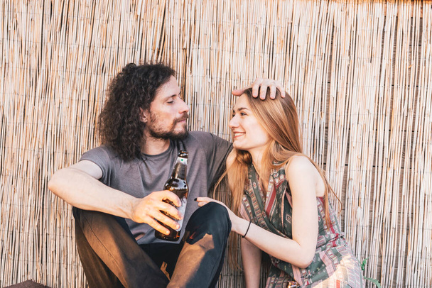 Young hipster couple sitting on the ground in a bar with afternoon light while drinking beer. Love and affection concept - Фото, изображение