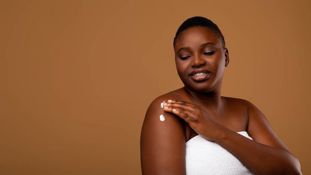 Όμορφη νεαρή μαύρη γυναίκα εφαρμογή ενυδατική κρέμα σώματος στους ώμους - Φωτογραφία, εικόνα