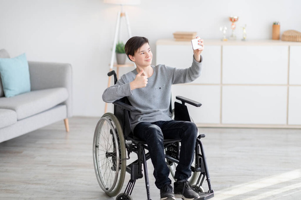 Joyeux adolescent handicapé en fauteuil roulant posant pour selfie, en utilisant un smartphone, montrant pouce vers le haut à la caméra à l'intérieur - Photo, image