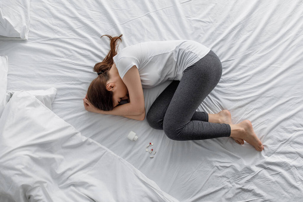 Droevige wanhoop jonge blanke vrouw die lijdt aan buikpijn liggend op bed met pot en verspreid pillen - Foto, afbeelding
