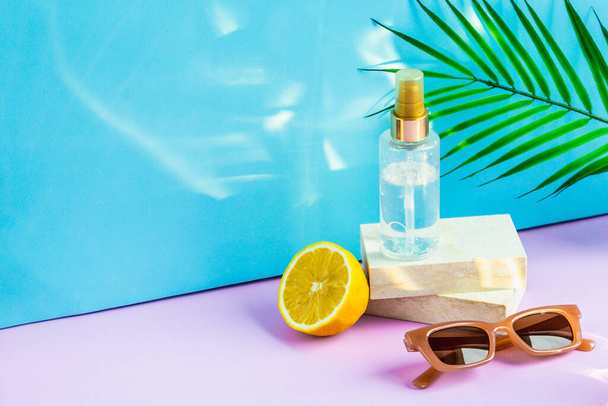 Cosmetics are on the podium on a summer background, in the rays of the sun. The spray is on the podium, half a lemon and sunglasses are lying next to it, and a palm leaf is hanging above it. - Fotografie, Obrázek