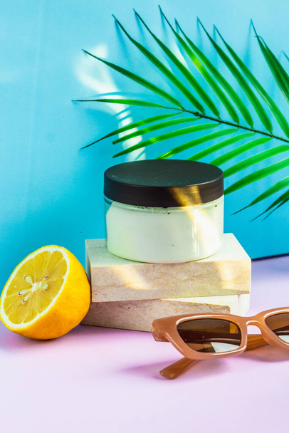 Cosmetics are on the podium on a summer background, in the rays of the sun. a jar of cream is on the podium, half a lemon and sunglasses are lying next to it, and a palm leaf is hanging above it. - Φωτογραφία, εικόνα